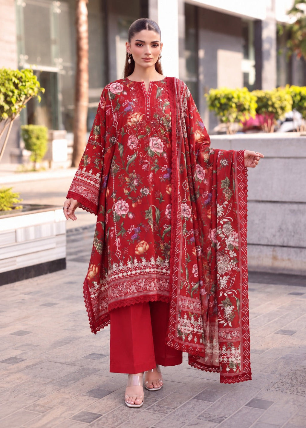 A woman modeling a red embroidered traditional outfit with a matching dupatta, standing outdoors on a paved walkway in front of a modern building.