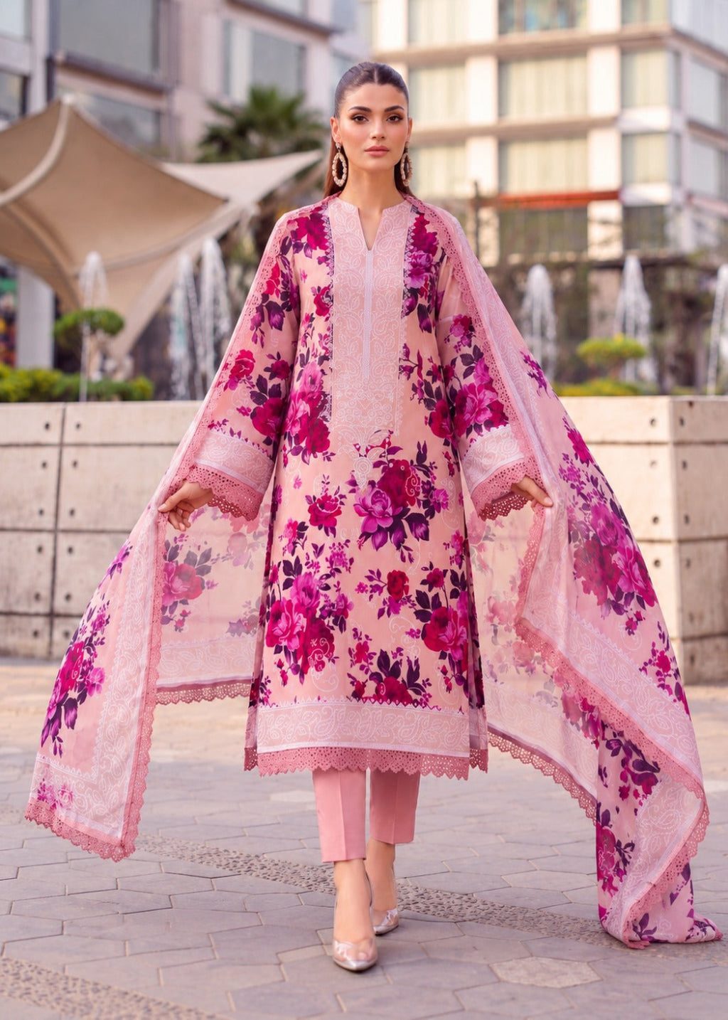 A woman models a three-piece pink ethnic suit with a vibrant magenta floral print, consisting of a long kurta, matching trousers, and a sheer dupatta, set against an outdoor urban background.