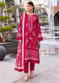 A woman models a three-piece traditional South Asian salwar kameez suit in a vibrant magenta pink, featuring detailed floral embroidery in gold, green, and white, paired with matching trousers and a coordinating dupatta, set against a blurred background of outdoor fountains.
