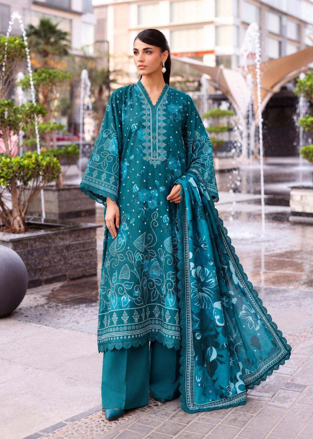 A woman stands outdoors in a plaza wearing a teal traditional outfit with matching pants and a sheer dupatta, featuring light blue floral embroidery and printed motifs.