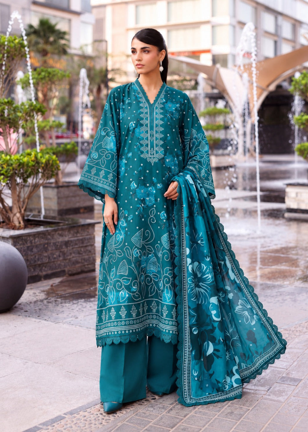 A woman stands outdoors in a plaza wearing a teal traditional outfit with matching pants and a sheer dupatta, featuring light blue floral embroidery and printed motifs.