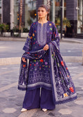 A woman models a three-piece traditional South Asian outfit in deep purple, featuring a kurta with colorful floral embroidery, matching wide-leg trousers, and a coordinating dupatta draped over her shoulder, set against a blurred outdoor urban background.