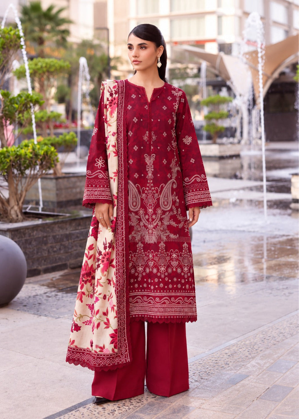 A woman models a three-piece traditional South Asian outfit in a park with water fountains. The outfit consists of a deep red kurta with intricate gold embroidery, matching red wide-leg trousers, and a cream dupatta with red floral patterns.