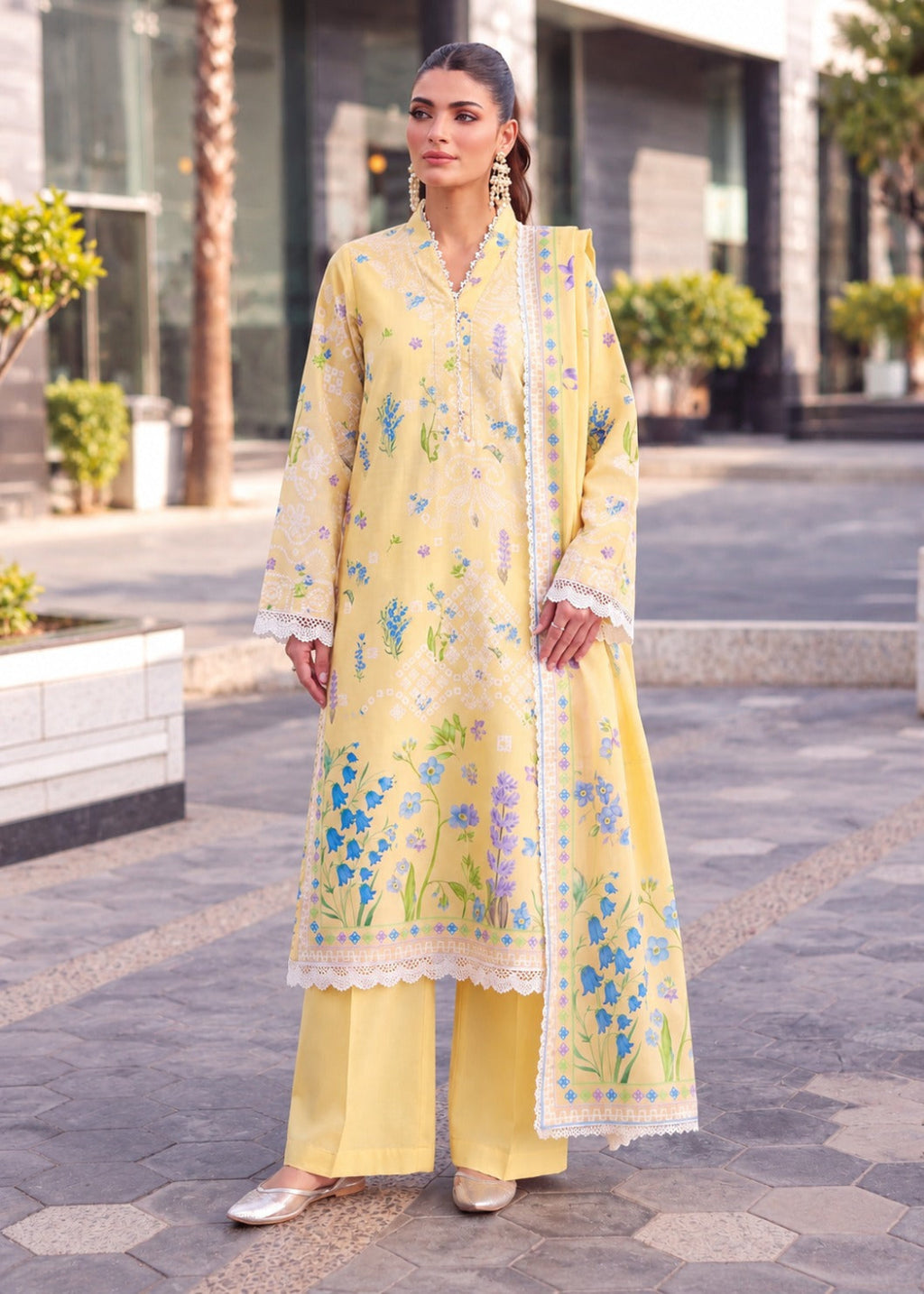 A woman models a light yellow three-piece traditional South Asian salwar suit set outdoors. The set includes a long kurta with blue and green floral embroidery and white lace trim, matching wide-leg trousers, and a coordinating dupatta.