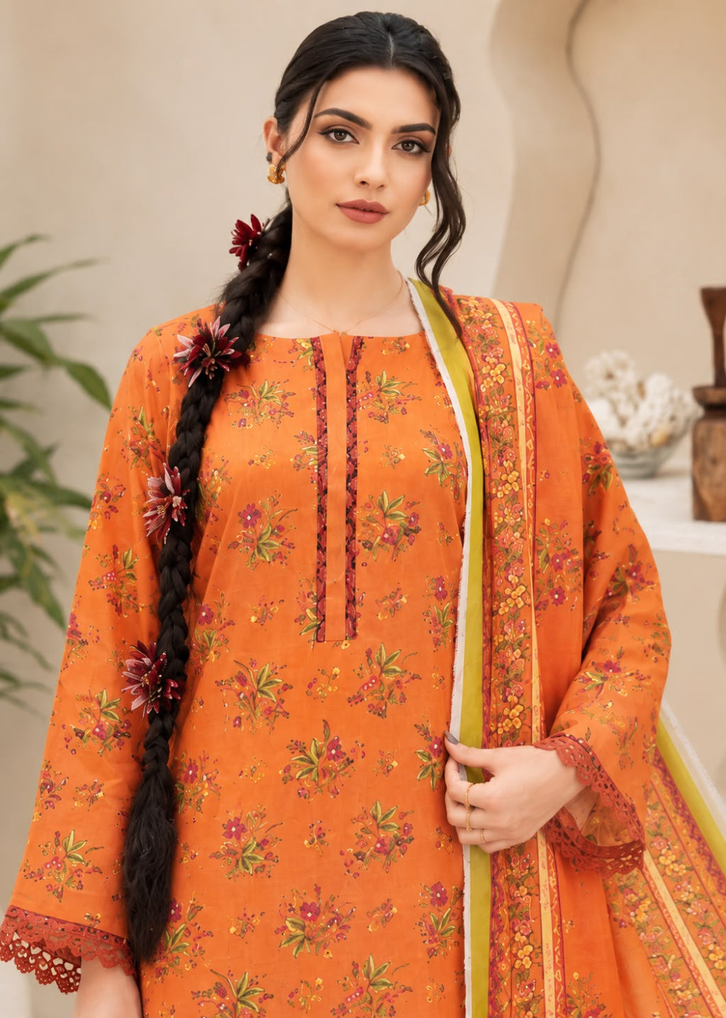 A woman modeling an orange floral salwar kameez suit with a matching dupatta. The kurta features a round neckline with vertical button placket, three-quarter sleeves with lace trim, and an allover floral print.