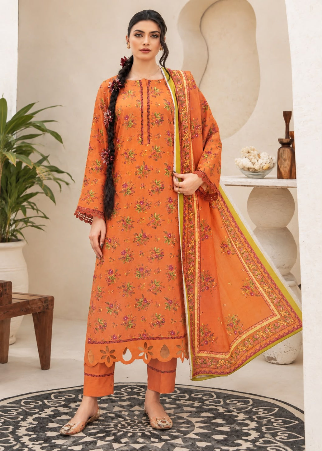 A woman models a traditional three-piece salwar kameez suit in orange, featuring floral embroidery, a matching dupatta, and cropped pants, set against a warm beige architectural background with a plant and decorative table.