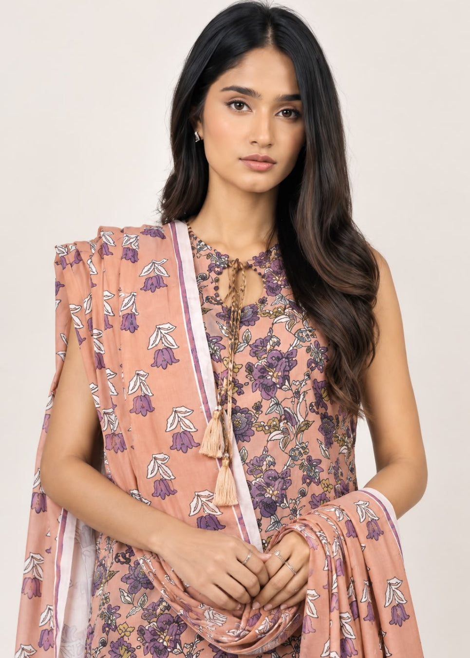 A woman modeling a peach-colored floral printed salwar suit set with a matching dupatta, featuring purple and white floral motifs, against a plain light beige background.