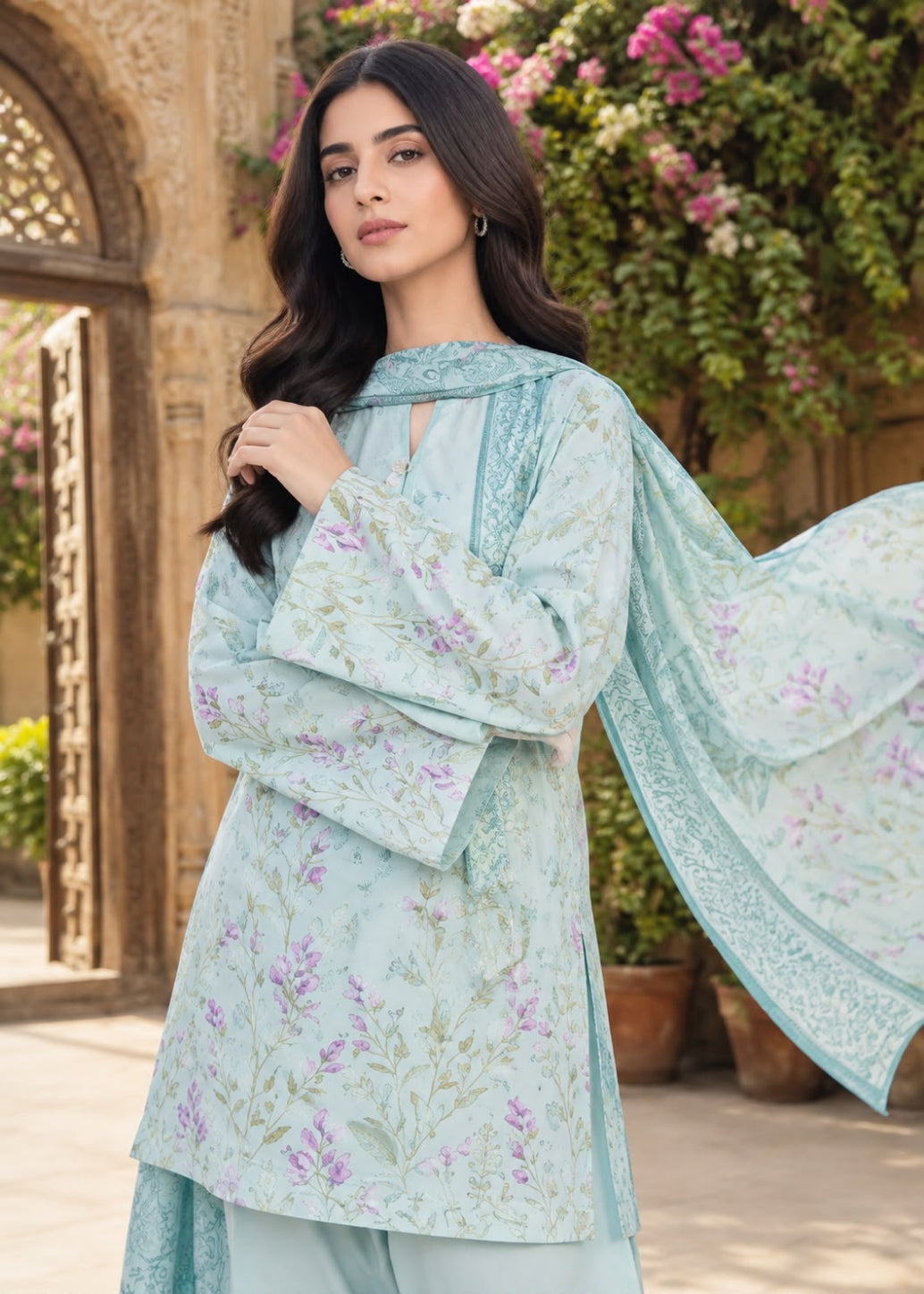 A woman modeling a light blue-green floral salwar kameez suit with a matching dupatta in an outdoor courtyard setting.