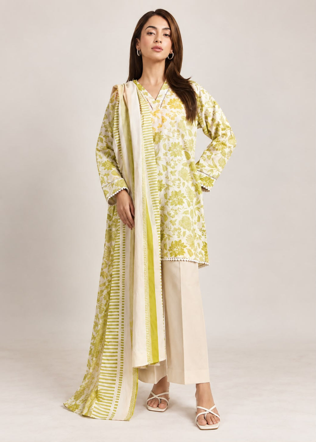 A woman models a traditional three-piece salwar kameez set in a studio setting. The outfit features a light yellow-green and white floral print kurta, beige wide-leg trousers, and a matching cream and yellow-green striped dupatta.