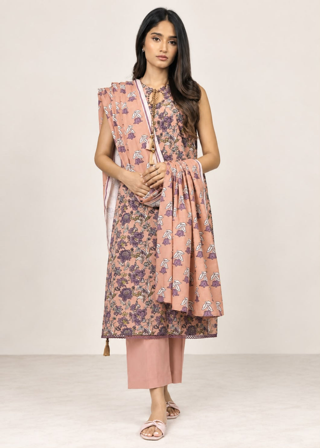 A woman models a traditional Indian outfit consisting of a sleeveless dusty pink kurta with a purple and white floral print, matching pink pants, and a coordinating dupatta, set against a plain beige studio background.