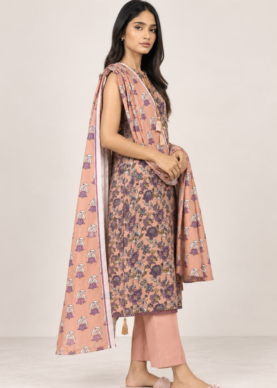 A woman modeling a peach-colored floral kurta set with matching printed dupatta and light pink pants against a plain beige studio background.
