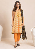 A woman models a traditional Indian outfit consisting of a sleeveless mustard yellow kurta with white floral patterns, orange cropped pants, and a black dupatta with yellow and gold geometric patterns, set against a warm beige background with decorative white ceramic vases.