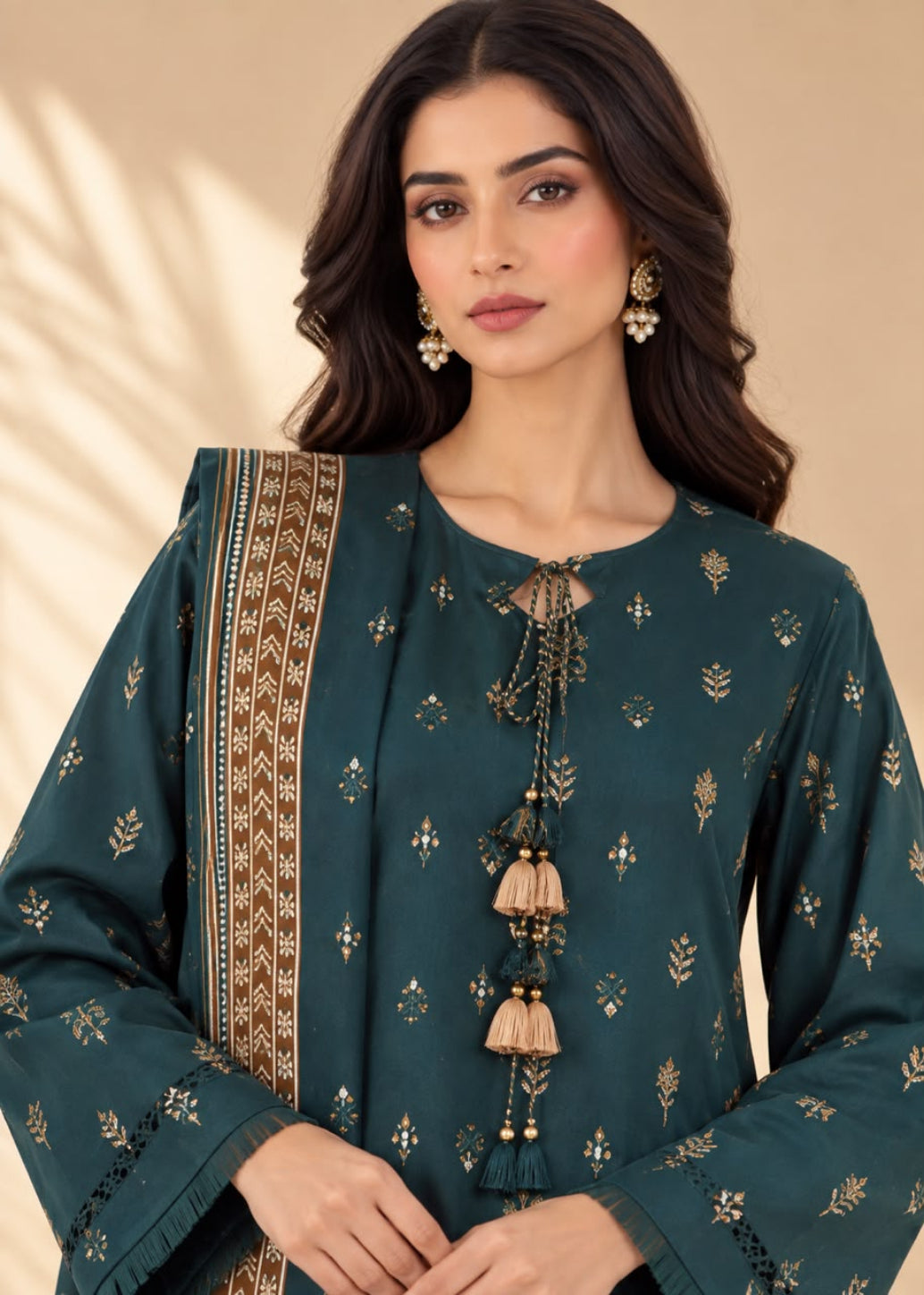A woman modeling a dark teal green kurta with gold floral prints, a matching patterned dupatta, and decorative tassel ties at the neckline, set against a warm beige background.