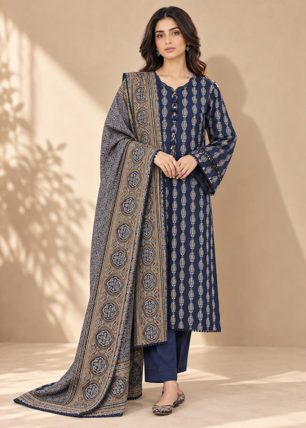 A woman models a navy blue three-piece salwar kameez suit with beige and white block prints, featuring a matching printed dupatta draped over her shoulder, set against a warm beige background with soft leaf shadows.