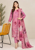 A woman models a three-piece pink ethnic suit set featuring a floral printed kurta, matching trousers, and a draped dupatta, standing next to a yellow woven chair against a neutral beige background.