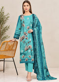 A woman models a teal blue floral printed salwar kameez suit with matching pants and a dupatta, standing next to a woven orange chair against a neutral beige background.