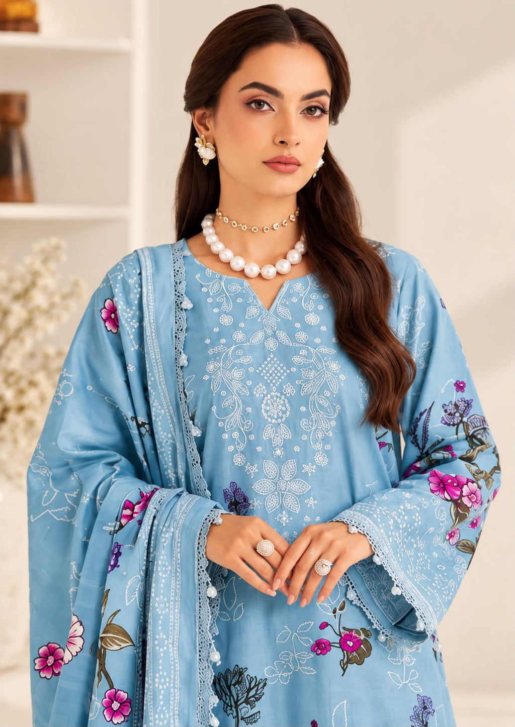 A woman modeling a light blue embroidered traditional outfit with floral print and a matching dupatta, styled with a pearl necklace and earrings in a bright indoor setting.