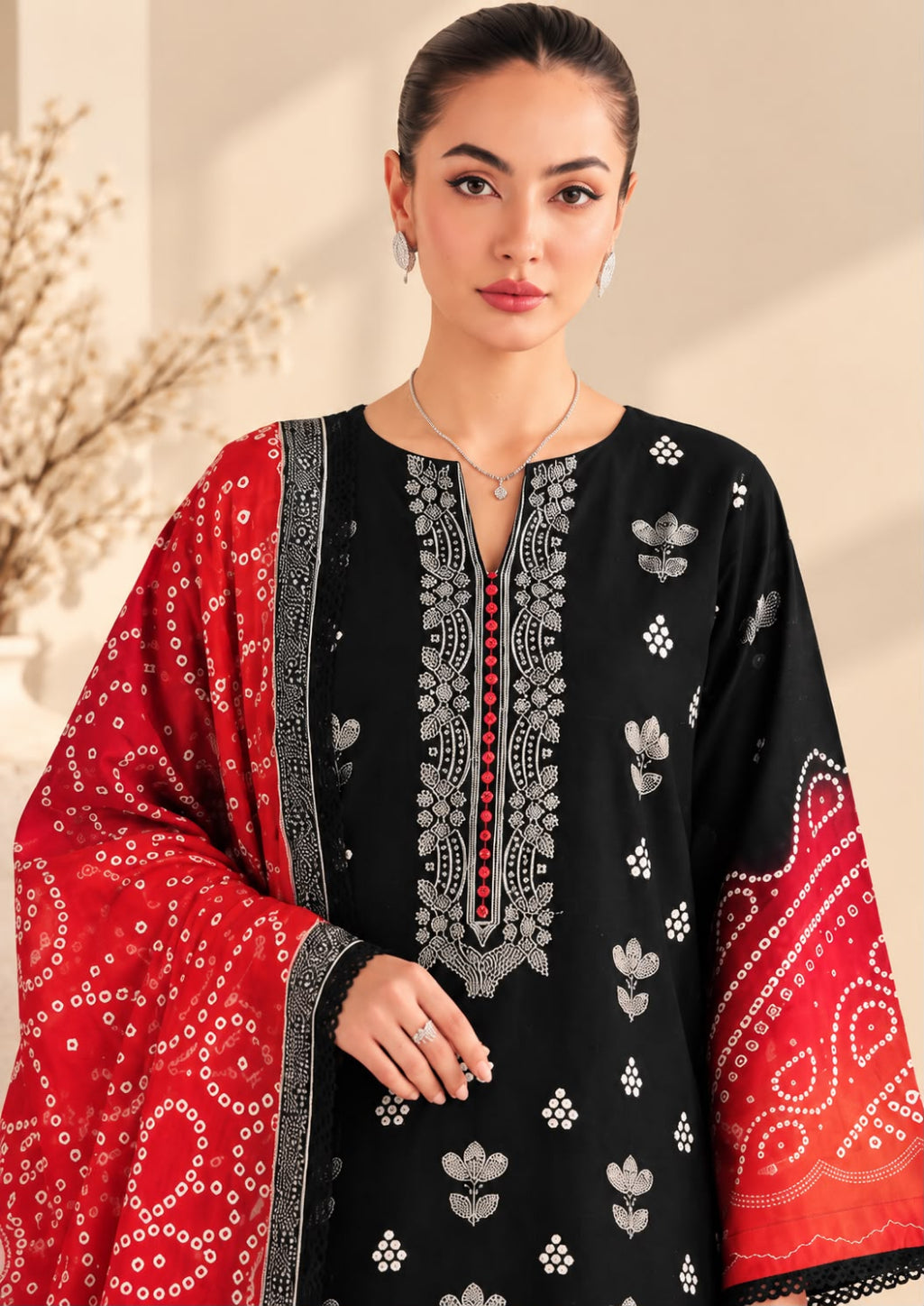 A woman modeling a black kurta with silver floral embroidery and a red dupatta featuring white bandhani patterns, set against a warm beige background.