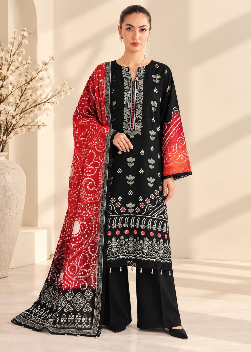 A woman models a black three-piece traditional outfit with red and white printed patterns, featuring a matching red dupatta draped over her shoulder, set against a warm beige background.