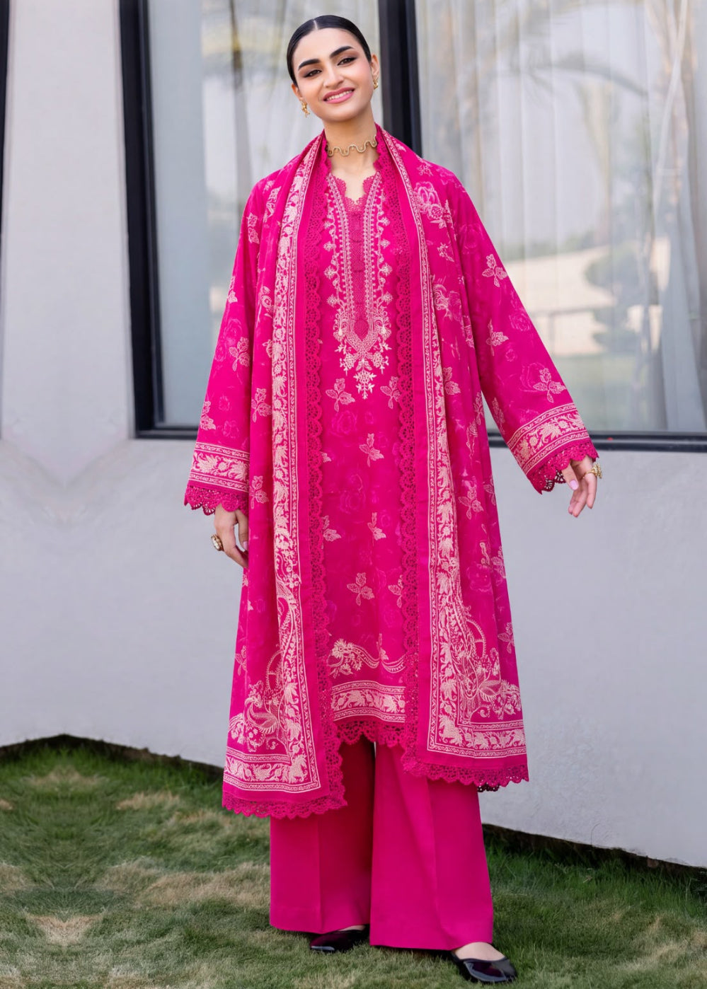 A woman modeling a bright pink traditional outfit with matching printed dupatta and wide-leg trousers, standing outdoors in front of a white wall and window.
