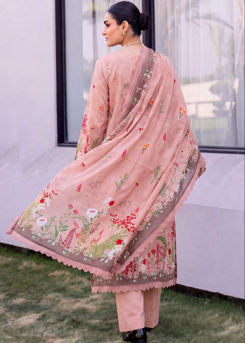 A woman modeling a peach-pink traditional outfit with floral embroidery and a matching dupatta, shown in a split front and back view outdoors near a building with large windows.