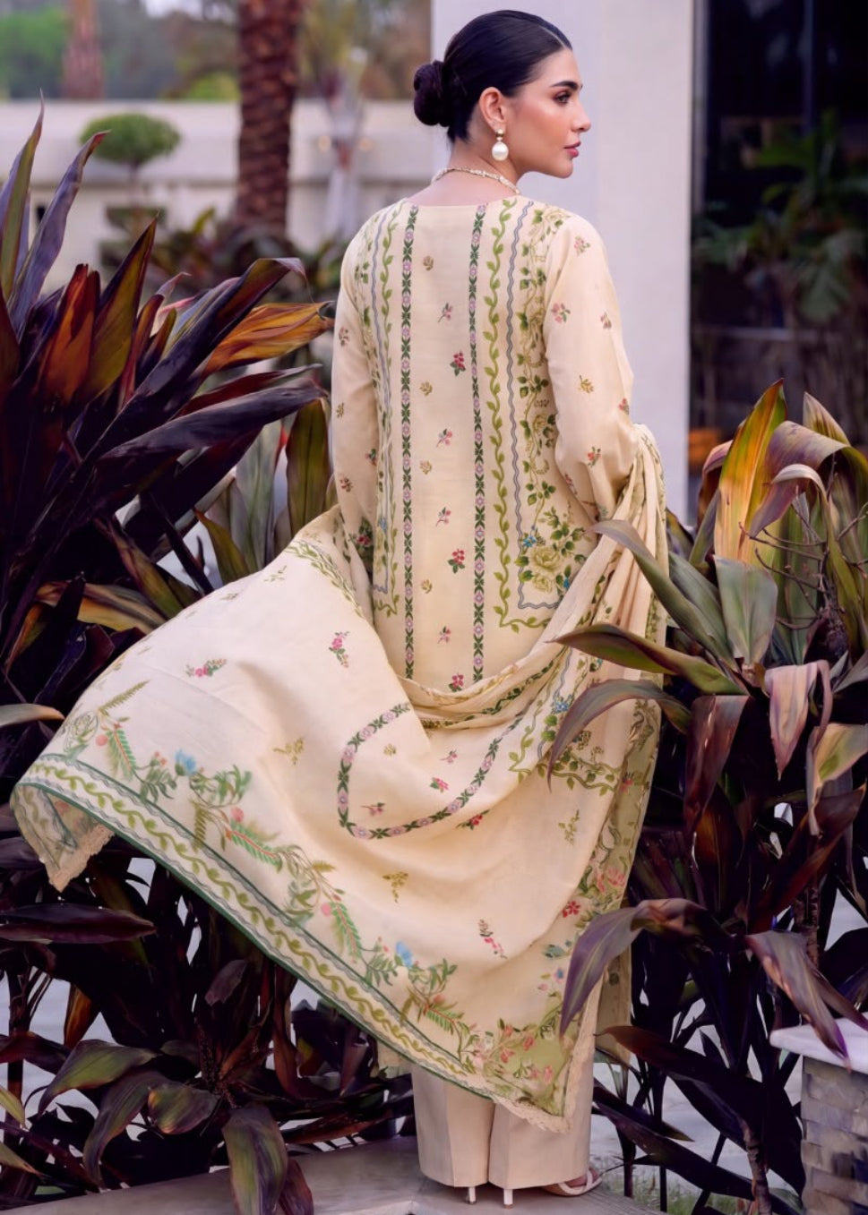 A woman modeling a traditional three-piece salwar kameez suit in an outdoor garden setting. The left panel shows her from the back, wearing a cream-colored tunic and matching pants with green floral embroidery and a green border.