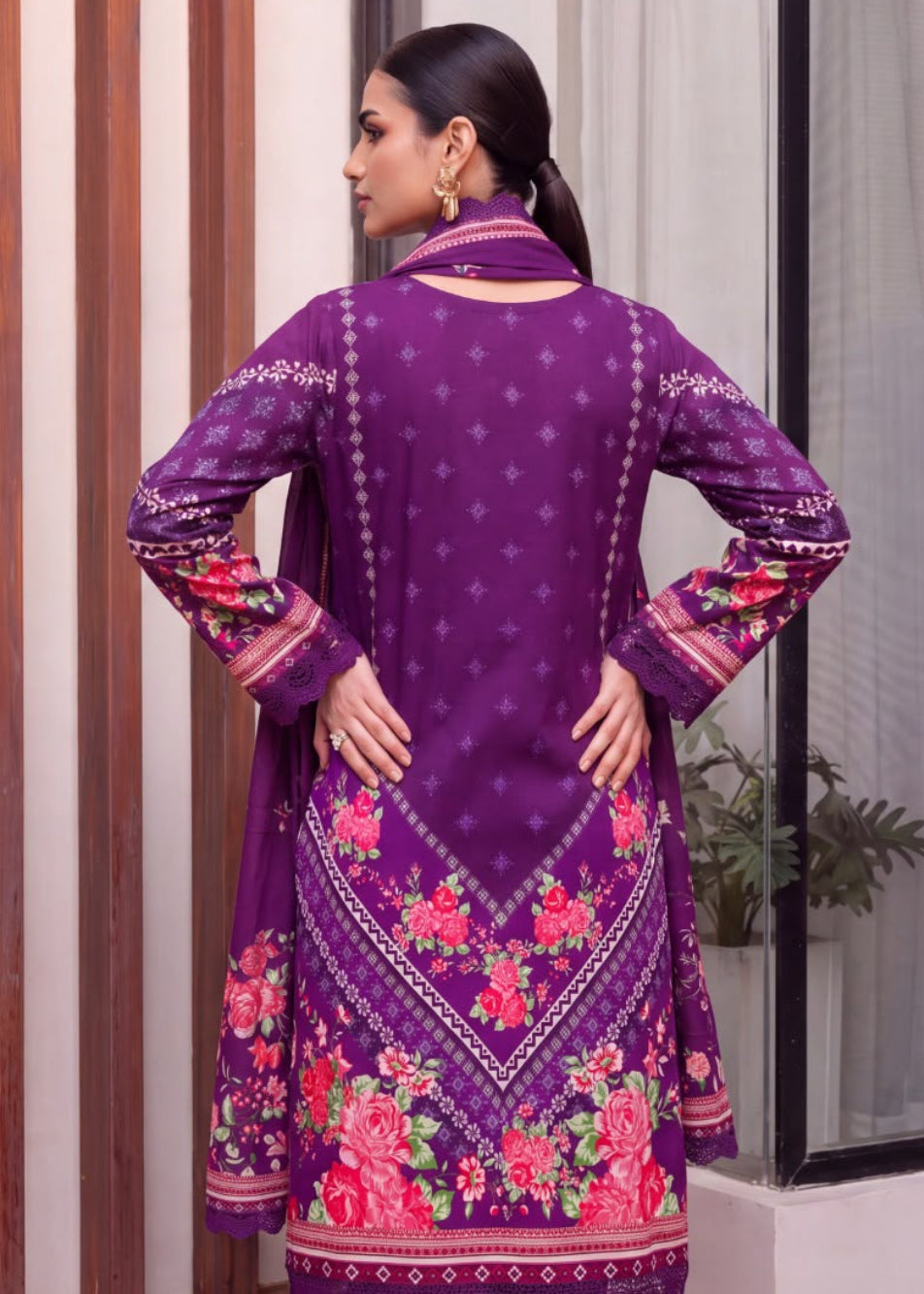 A woman modeling a purple traditional outfit with floral embroidery, shown in a split view: one side shows the back of the kurta with embroidered sleeves and hem, and the other shows a close-up of the draped fabric and face. The setting appears to be an indoor architectural space with vertical wood-toned panels.