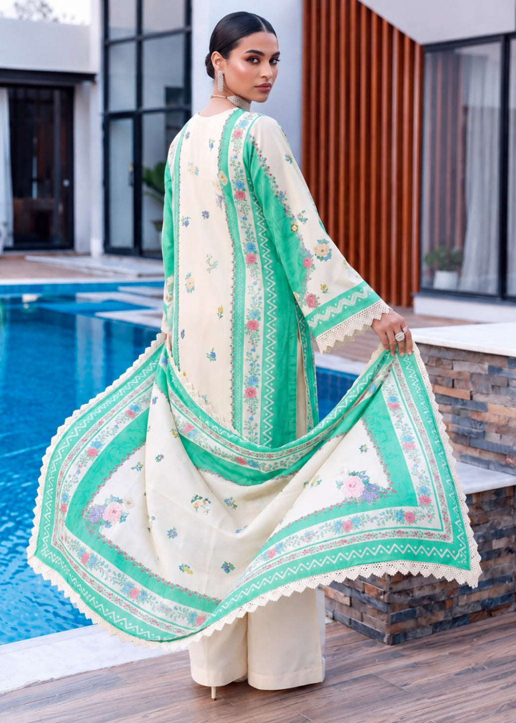 A woman stands by a swimming pool wearing a cream-colored traditional outfit with mint green borders and floral prints. She is shown from the back, holding out the wide sleeves and dupatta to display the pattern and color details.