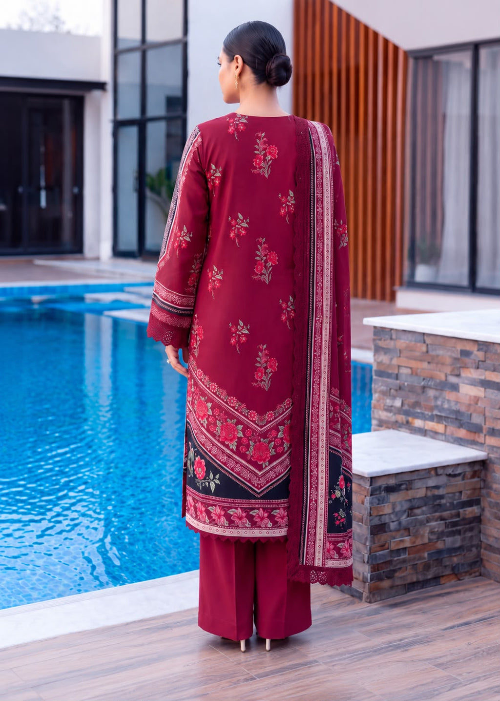 A woman stands with her back to the camera wearing a deep red three-piece traditional outfit with floral embroidery and a matching dupatta, positioned next to a swimming pool in a modern outdoor setting.