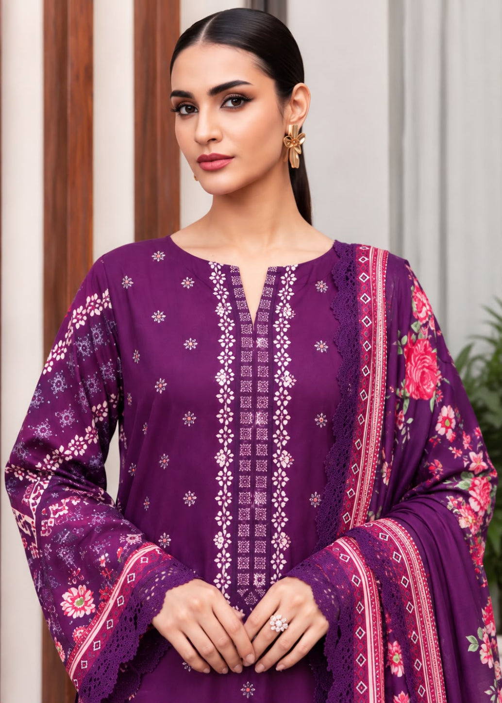 A woman modeling a purple three-piece ethnic suit with intricate white embroidery, floral prints, and a matching dupatta, set against a neutral indoor background.