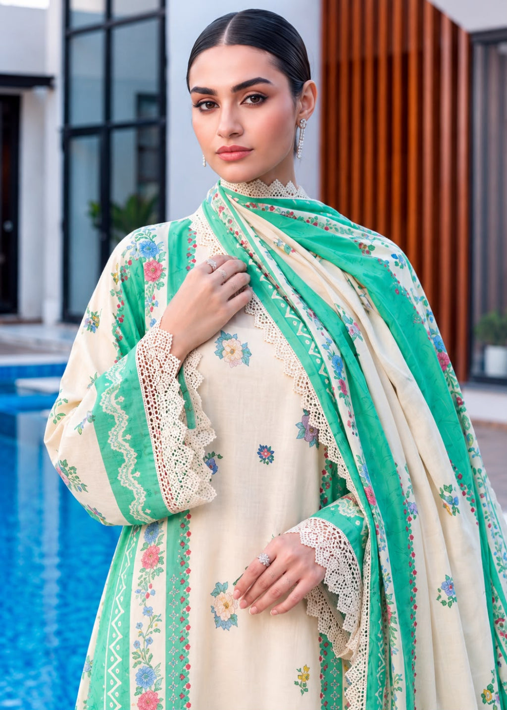 A woman models a traditional South Asian outfit featuring a cream-colored kurta with delicate floral embroidery, a matching green dupatta with lace borders, and a green embroidered jacket. The background shows a modern outdoor setting with a swimming pool on the left and a wooden slatted wall on the right.