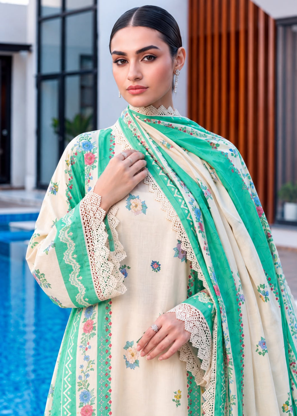 A woman models a traditional South Asian outfit featuring a cream-colored kurta with delicate floral embroidery, a matching green dupatta with lace borders, and a green embroidered jacket. The background shows a modern outdoor setting with a swimming pool on the left and a wooden slatted wall on the right.