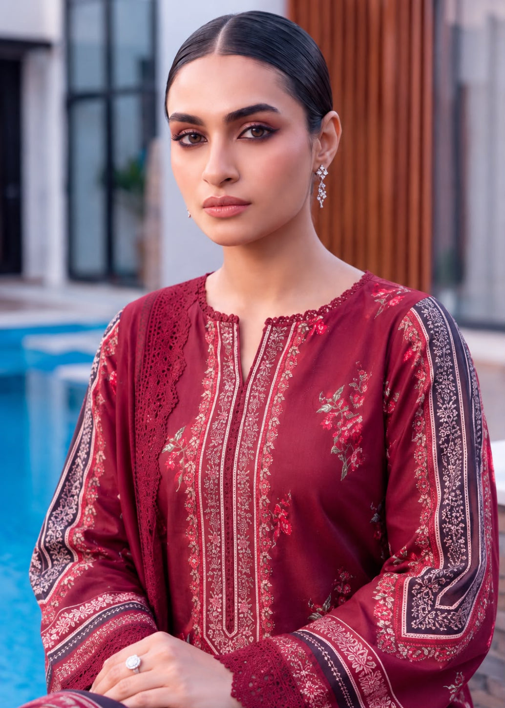A woman wearing a deep maroon traditional embroidered kurta with intricate floral and geometric patterns, posed in front of a modern building and pool background.