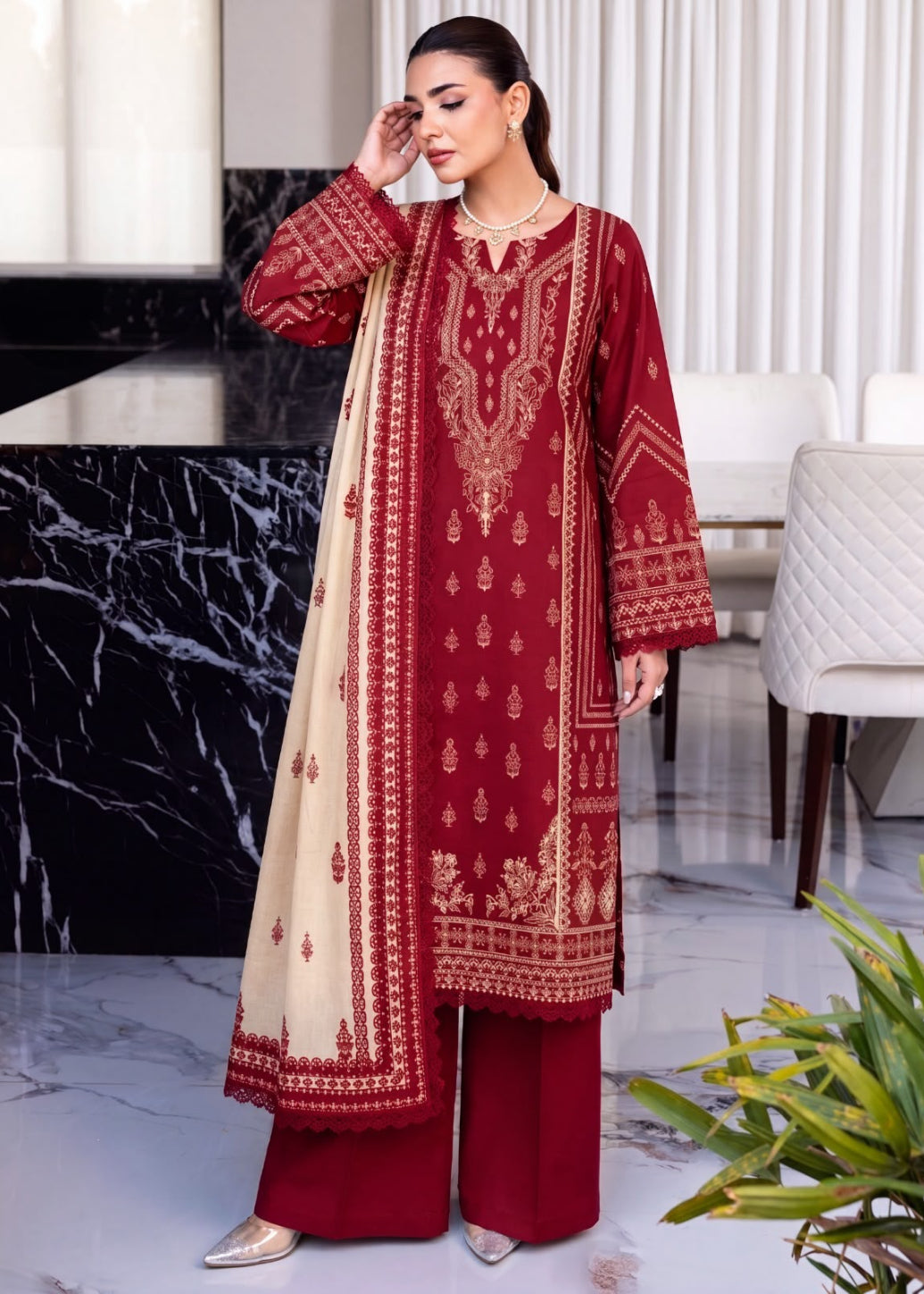 A woman models a traditional three-piece Indian outfit consisting of a deep red kurta with gold embroidery, matching red trousers, and a beige dupatta with red borders, set against a modern interior background.