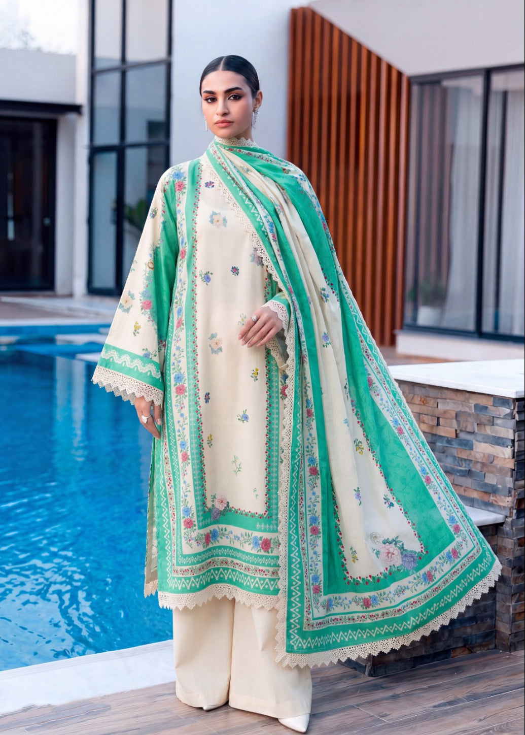 A woman models a traditional South Asian outfit consisting of a cream-colored kurta with colorful floral embroidery, matching cream trousers, and a vibrant green dupatta with intricate borders, standing next to a swimming pool.