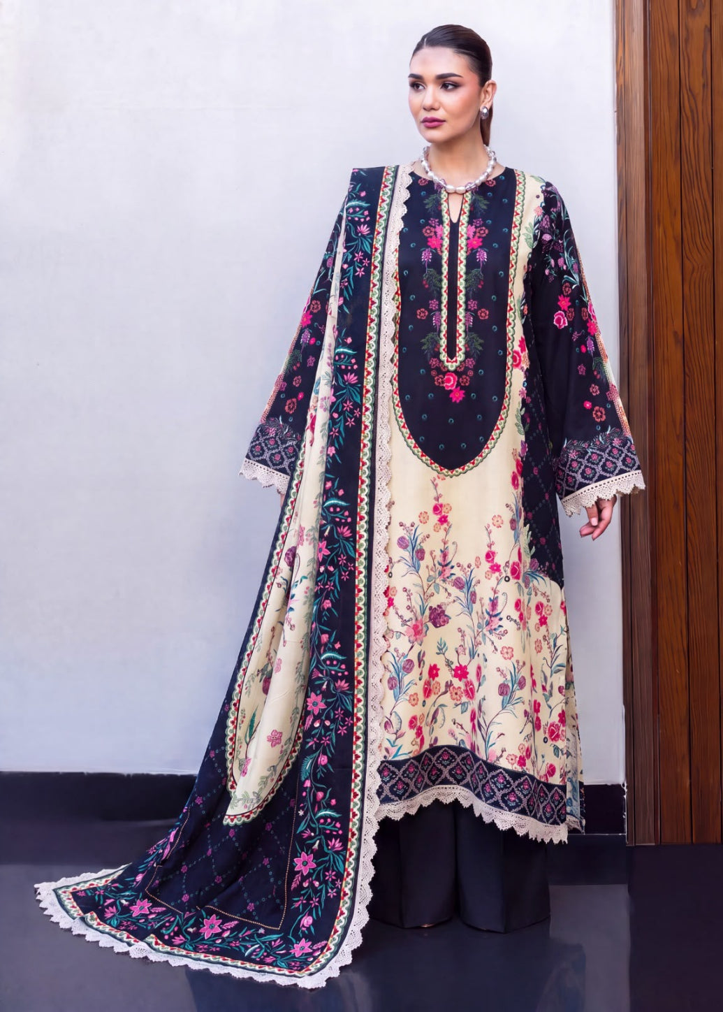 A woman models a traditional South Asian salwar kameez suit with a floral print. The outfit features a cream-colored kurta with pink and green floral embroidery, a navy blue yoke with gold embroidery, and a matching navy blue dupatta with white lace borders and pink floral prints.