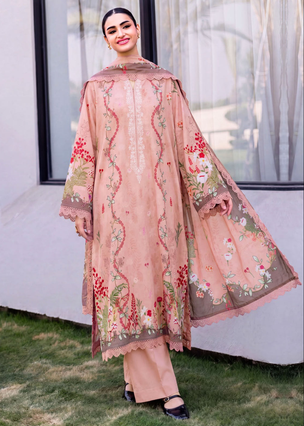 A woman models a traditional three-piece salwar kameez suit in dusty pink, featuring intricate floral embroidery in red, green, and gold, paired with matching wide-leg trousers and a dupatta, set against an outdoor backdrop of a white wall and green grass.