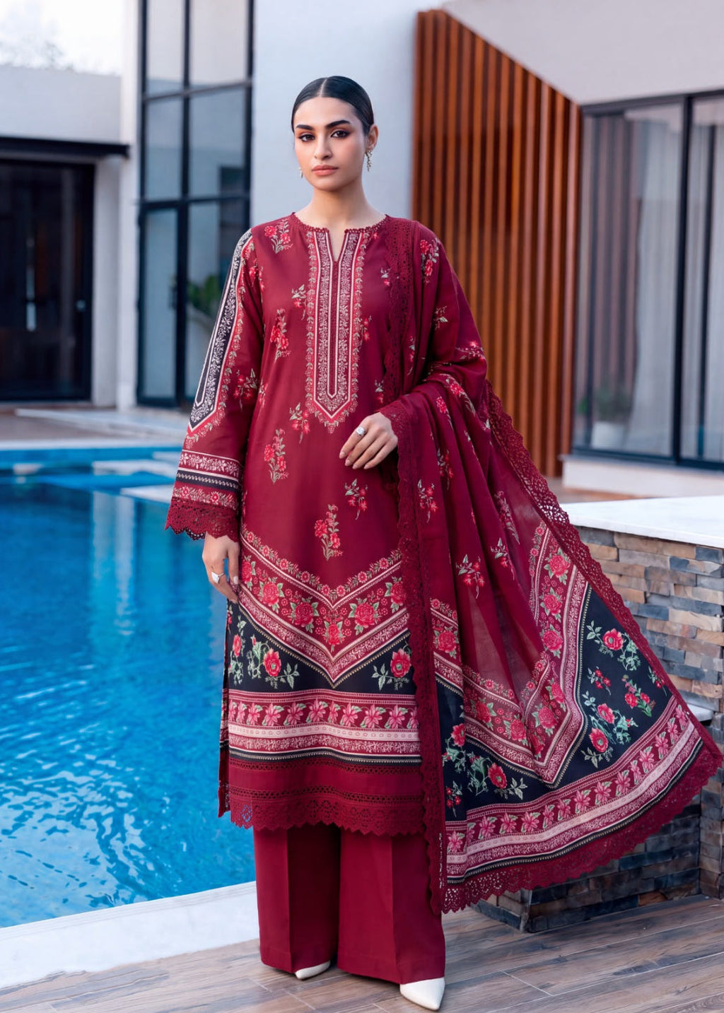 A woman models a three-piece traditional South Asian outfit consisting of a deep red embroidered kurta, matching trousers, and a sheer red dupatta with a black border and floral print, set against a modern poolside background.