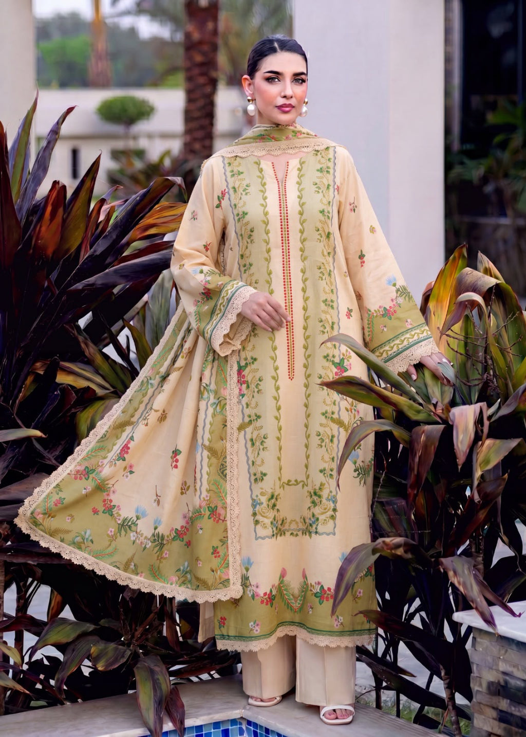 A woman models a traditional three-piece salwar kameez suit in a beige and olive green colorway with intricate floral embroidery, set against a background of tropical plants.