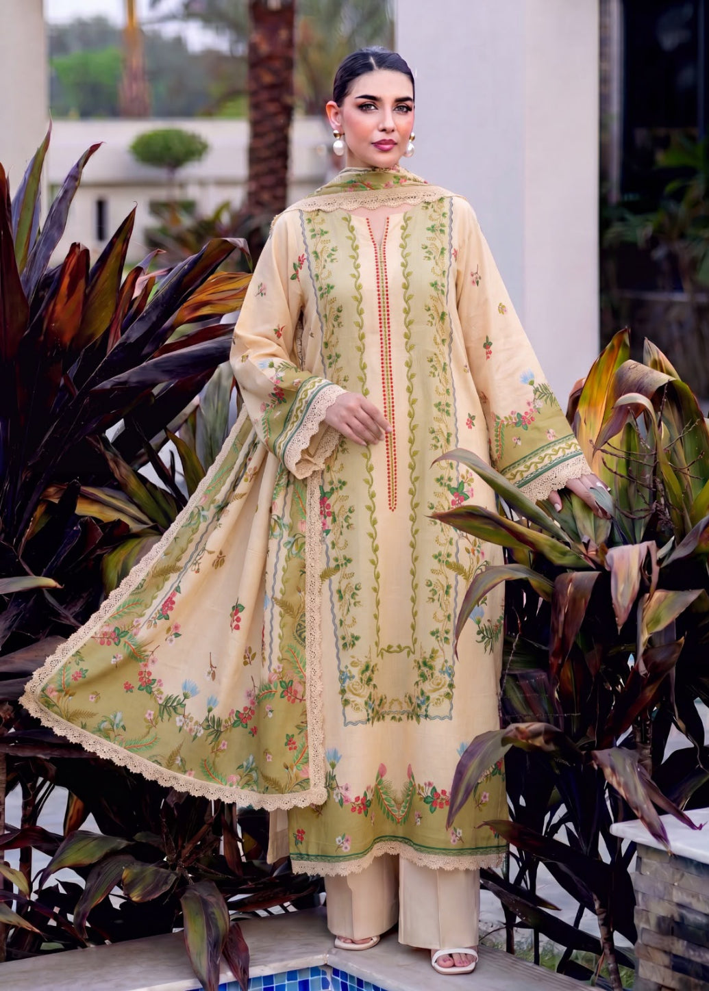 A woman models a traditional three-piece salwar kameez suit in a beige and olive green colorway with intricate floral embroidery, set against a background of tropical plants.