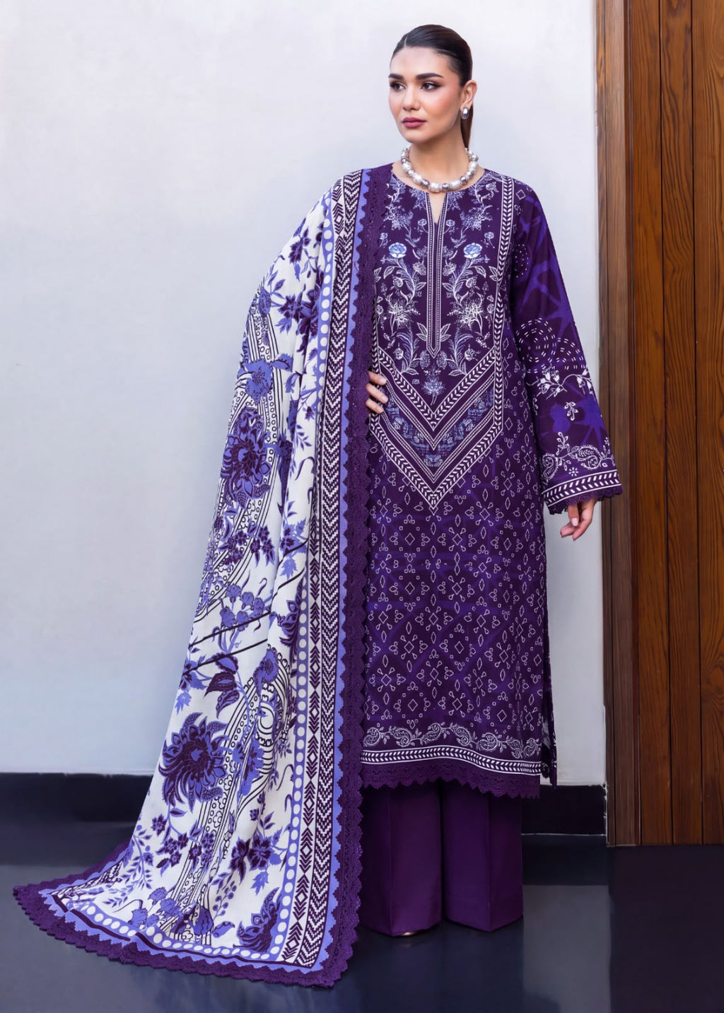 A woman models a traditional South Asian three-piece outfit consisting of a purple embroidered kurta, matching purple trousers, and a white dupatta with purple floral patterns and borders, set against a neutral indoor background.