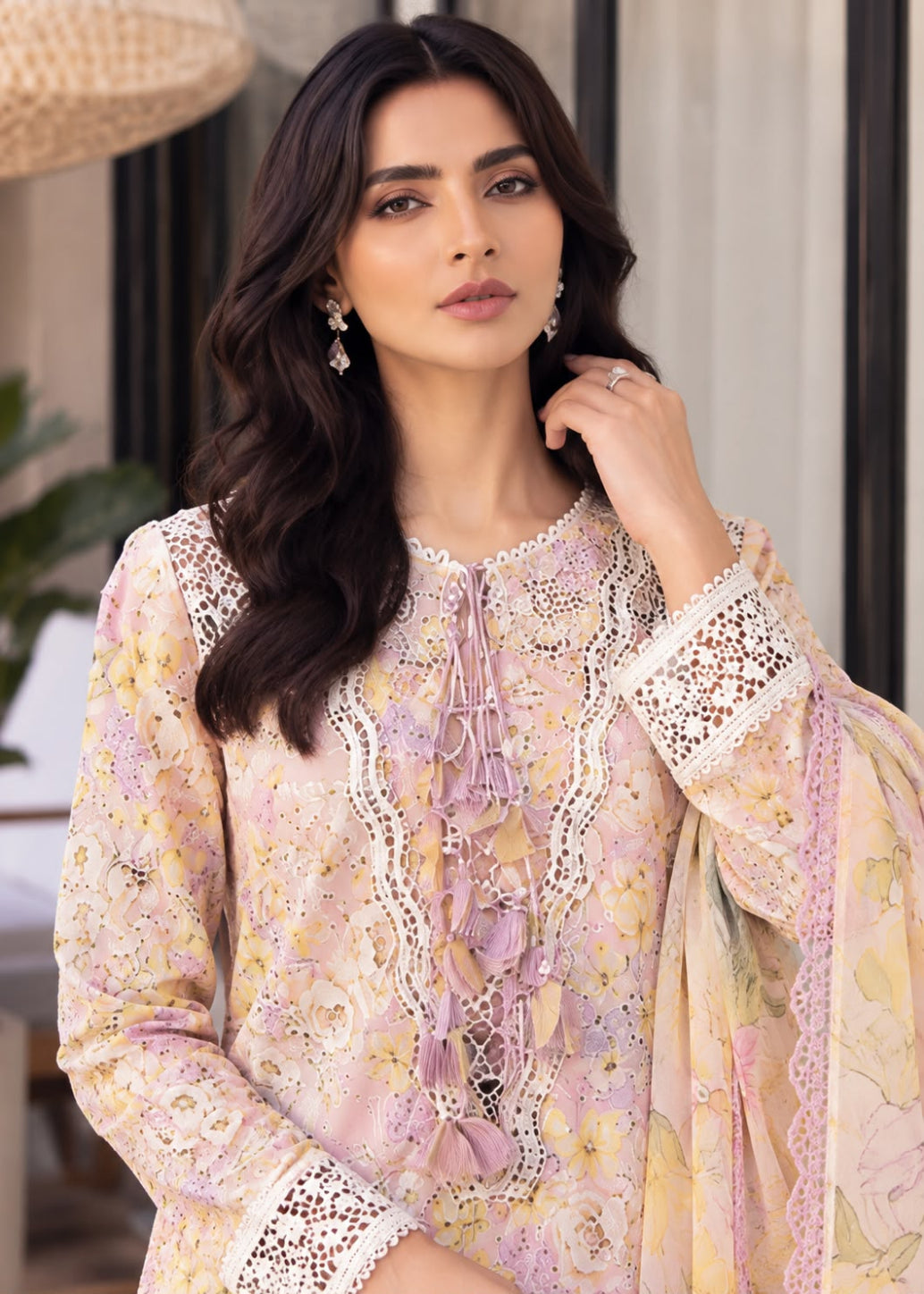 A woman wearing a pastel pink and yellow floral printed traditional outfit with intricate lace embroidery and a matching dupatta, posed in a softly lit indoor setting.