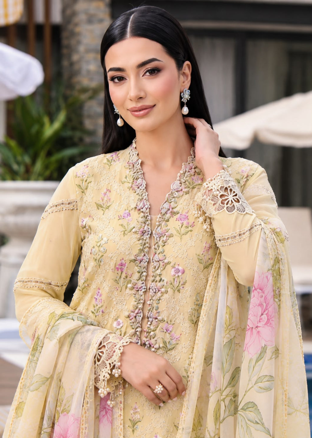 A woman wearing a traditional embroidered beige and pale yellow ethnic outfit with pink floral prints, lace cuffs, and a matching dupatta, photographed in an outdoor resort-like setting.