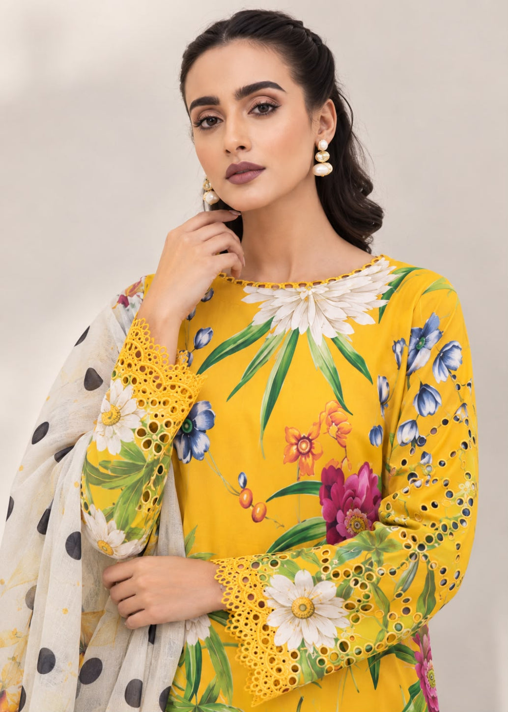 A woman models a bright yellow three-piece salwar kameez suit with a colorful floral print, featuring a matching yellow kameez top, printed trousers, and a white dupatta with black polka dots.