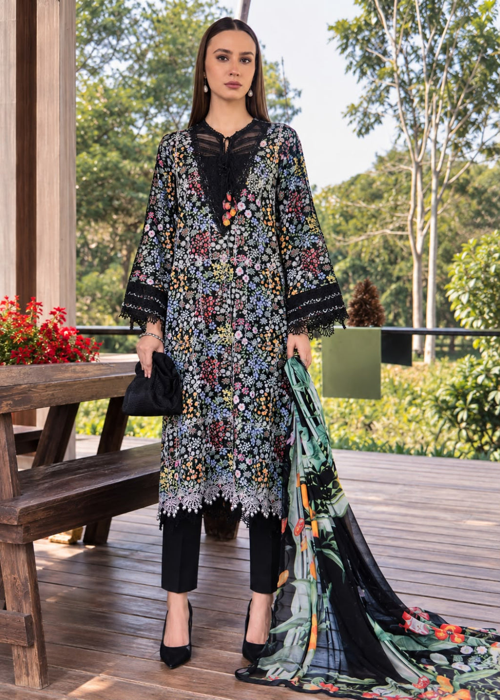 A woman stands on a wooden deck wearing a black embroidered tunic with multicolor floral patterns, black pants, and a matching printed dupatta. She is posed outdoors with trees and a bench in the background.