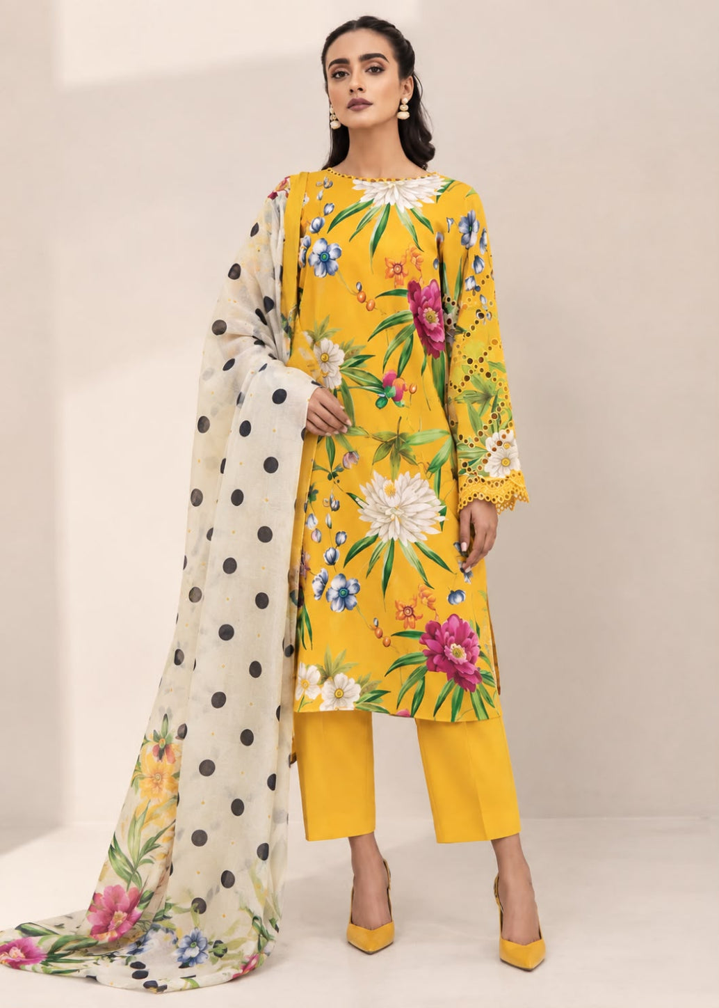 A woman models a three-piece traditional outfit featuring a bright yellow kurta with a colorful floral print, matching yellow trousers, and a cream dupatta with black polka dots and floral accents, set against a neutral beige studio background.