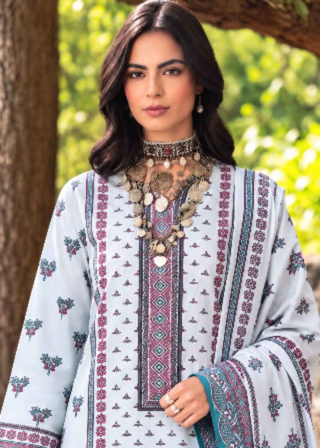 A woman wearing a light blue traditional embroidered kurta with floral and geometric patterns, styled with a chunky coin necklace and earrings, set against a soft-focus outdoor background of trees.
