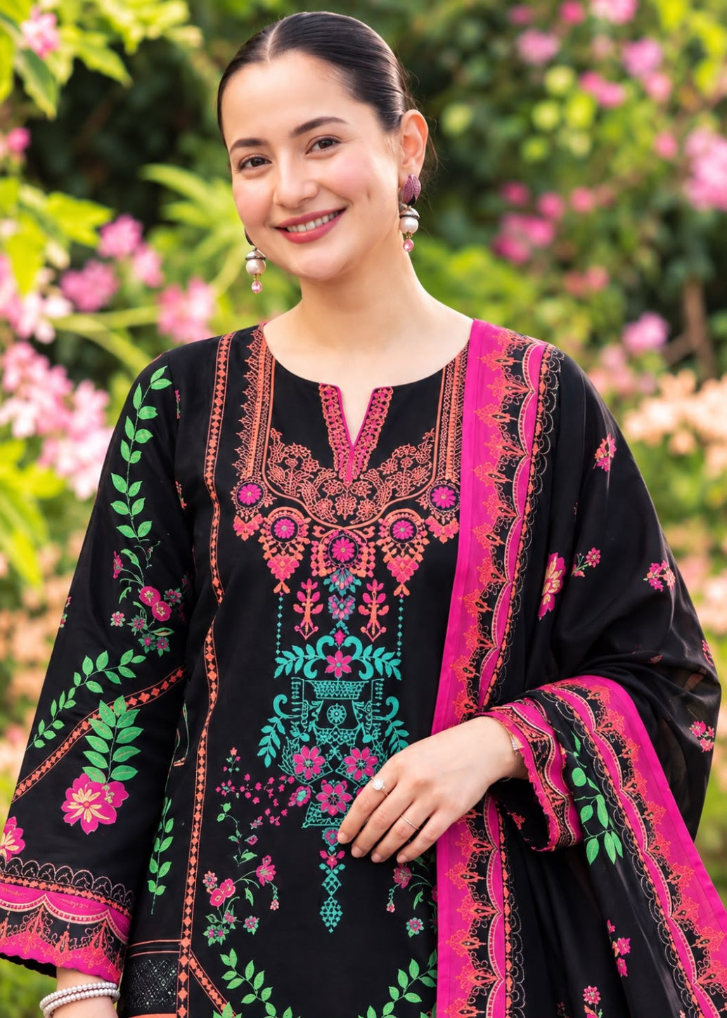 A woman wearing a black embroidered traditional outfit with bright pink, green, and orange details, standing outdoors in front of a blurred garden background of green foliage and pink flowers.