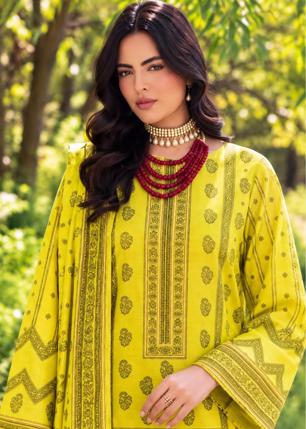 A woman wearing a bright yellow traditional tunic with dark green printed patterns and a matching dupatta, styled with layered red beaded necklaces, set against a soft-focus green outdoor background.