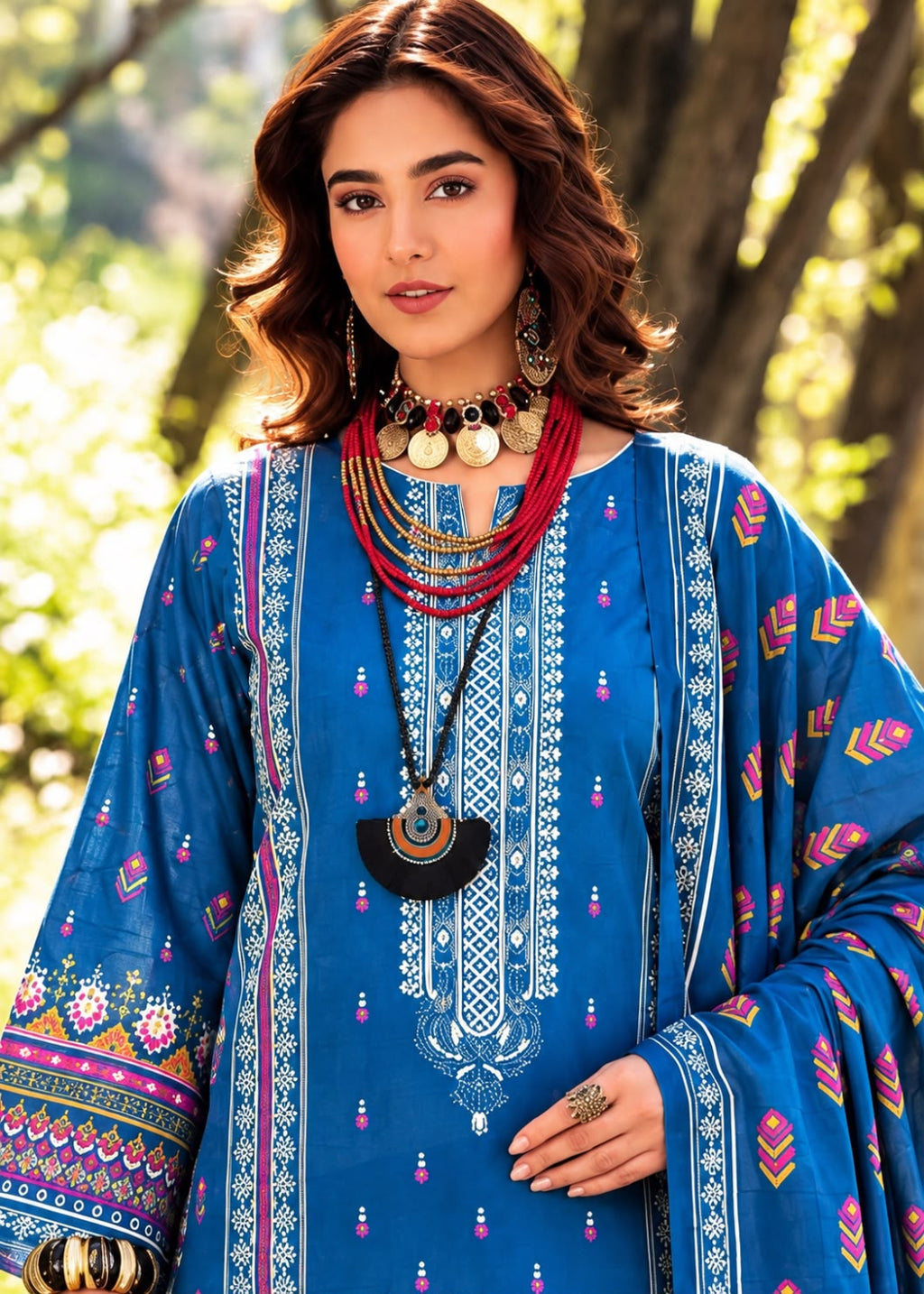 A woman wearing a blue embroidered traditional outfit with colorful geometric patterns, layered necklaces, and earrings, photographed outdoors against a blurred tree background.